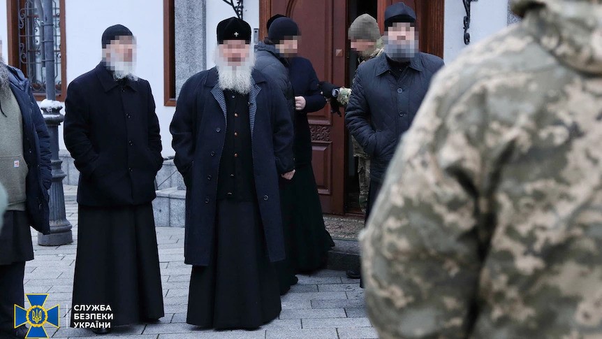 Three Orthodo priests with their faces blurred stand surrounded by military. 