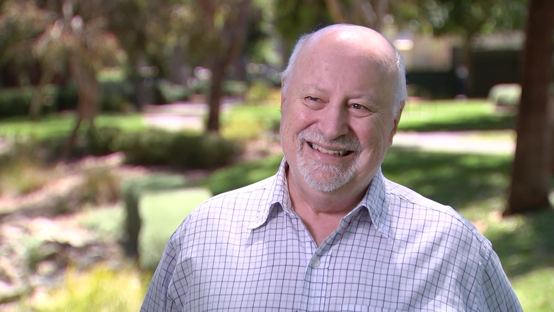 A smiling man with white hair and a white beard stands with trees and grass behind him