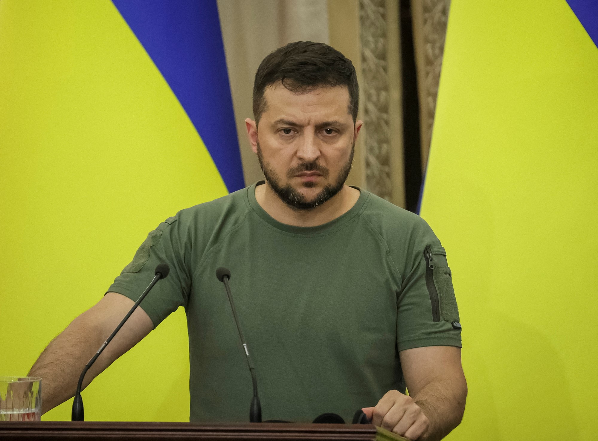 Volodymyr Zelenskyy stands behind a lecturn in front of Ukrainian flags.