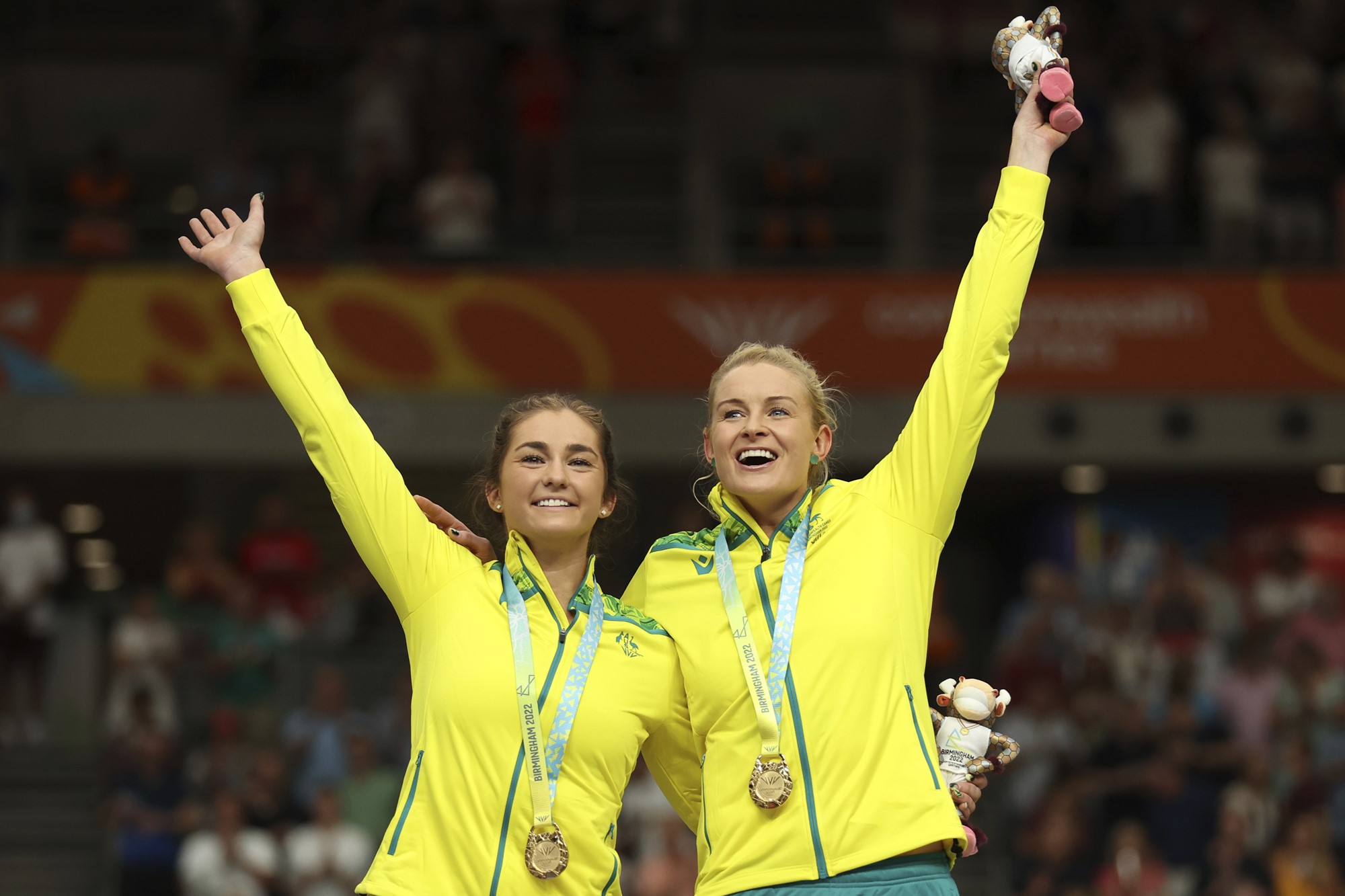 Two Australian athletes smile and wave gold medals on a podium