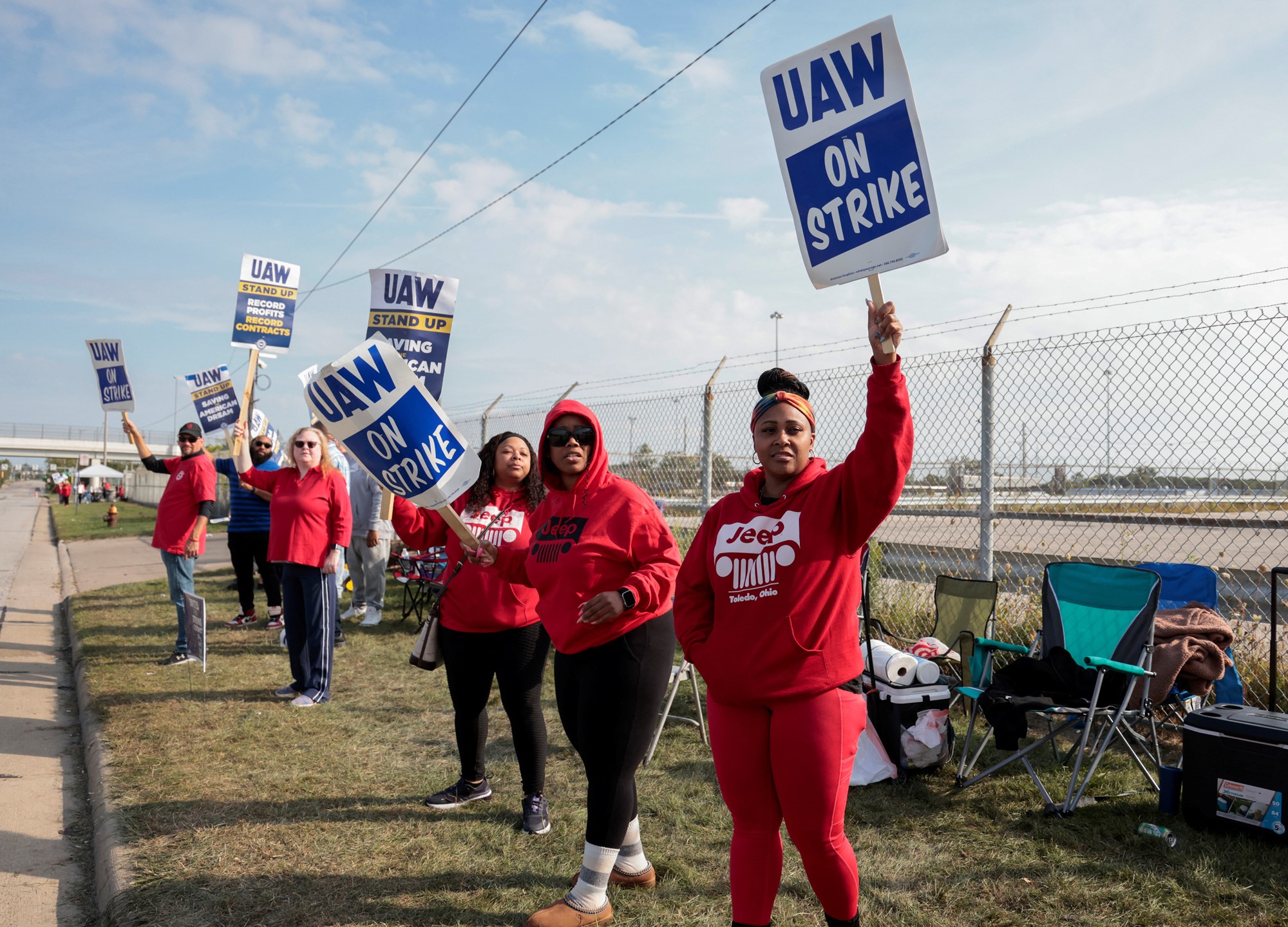 Workers wearing red hoodies and shirts hold picket signs outside a plant.