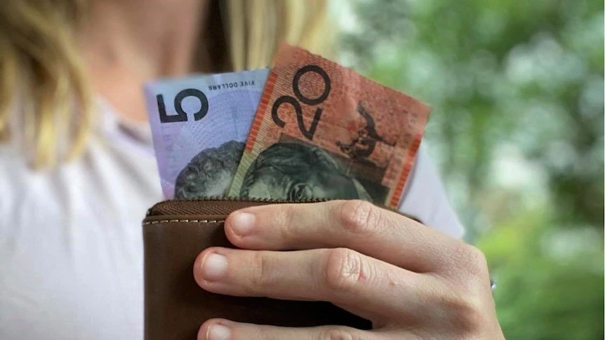 A person with short blonde hair holding a wallet with $5 and $20 notes pointing out of it