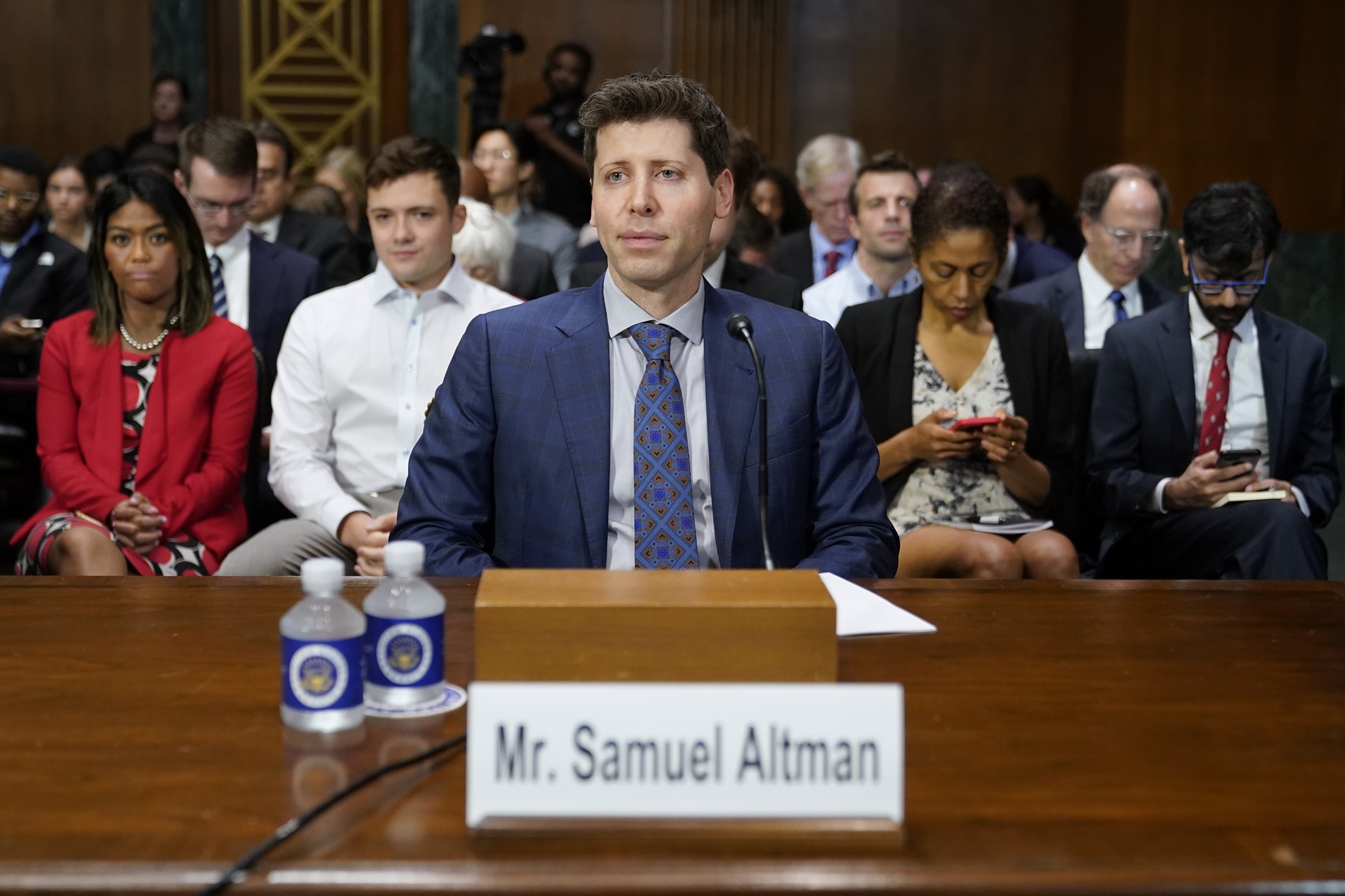 A white man in a suit sits at a bench in US Congress, with people behind him, and speaks into a microphone. Water and his name tage on the desk in front of him