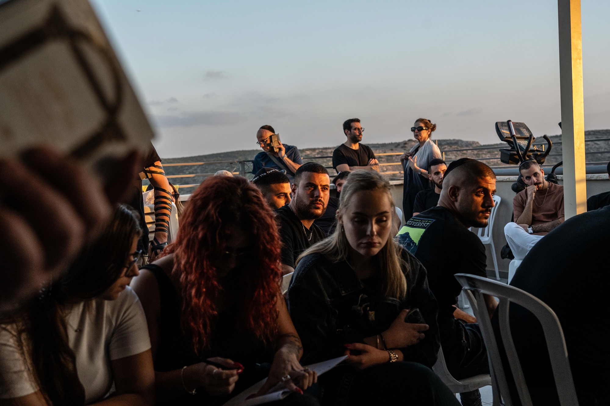 A group of people sit on chairs, mourning, as the sun sets behind them