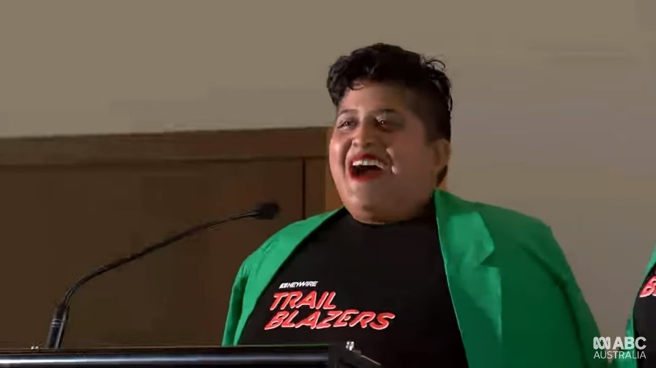 A young woman with short hair and a green blazer wearing a Trail Blazers shirt speaks by a podium.