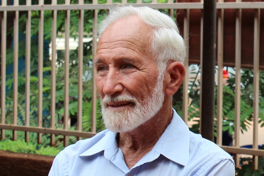 An older white man with white hair stands in front of a barred fence.