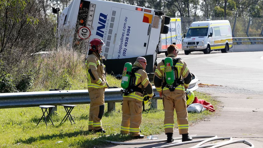 Three firefighters stand on the side of a road near an overturned bus