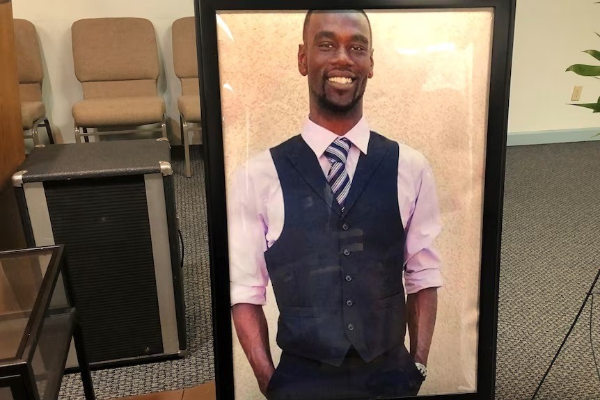 A framed photo of a Black man wearing a waistcoat and lilac shirt and tie with his hands in his pockets.