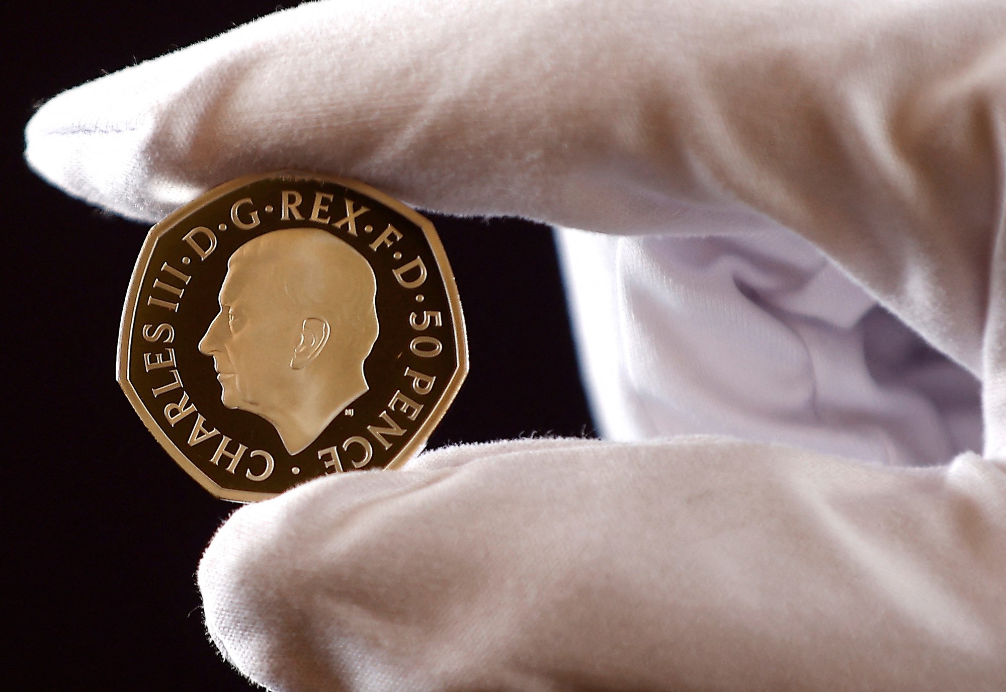 A coin featuring the side image of King Charles is held between a thumb and index finger.