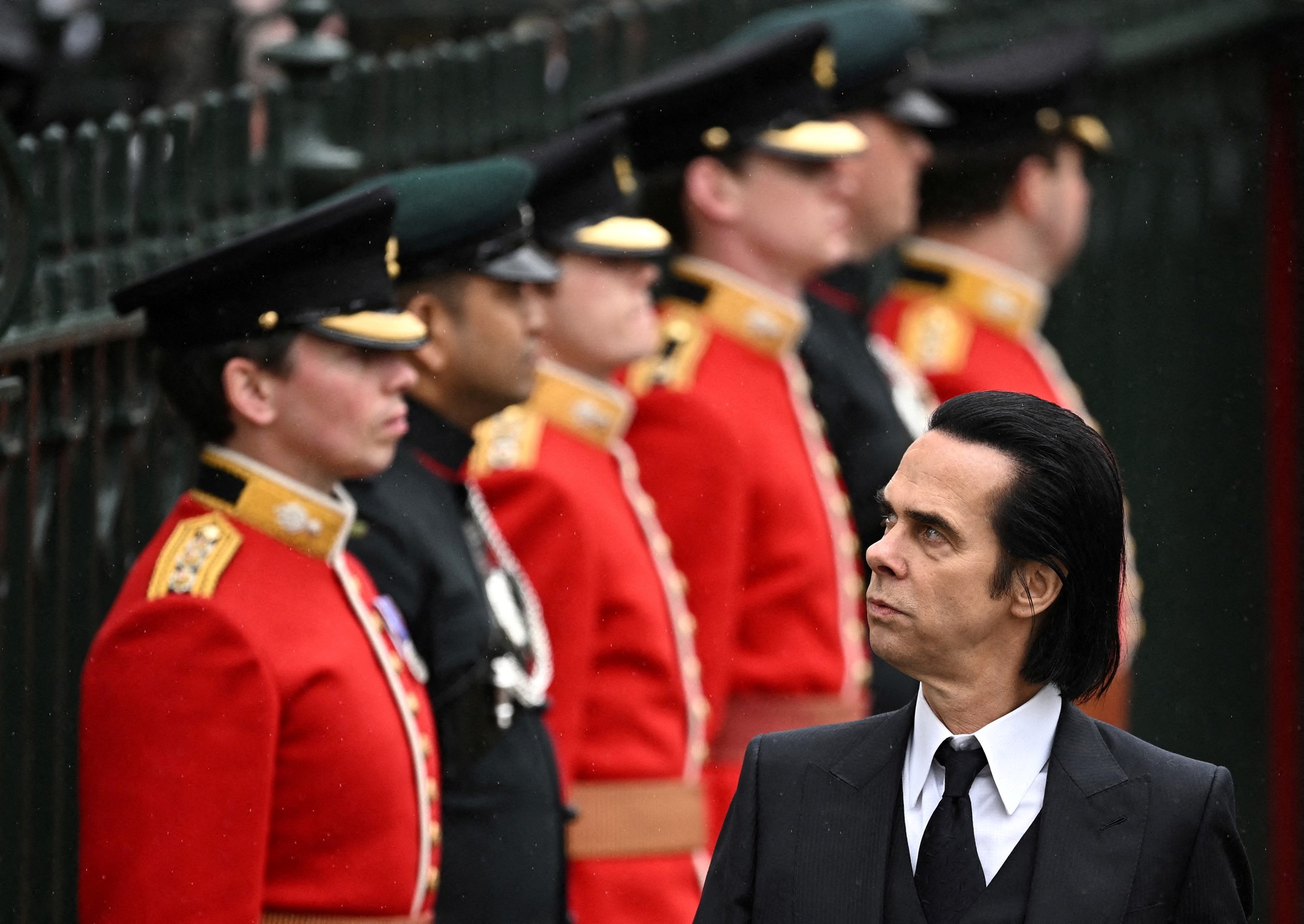 Nick Cave, in the foreground, looks to the side where a row of troops stand behind him,