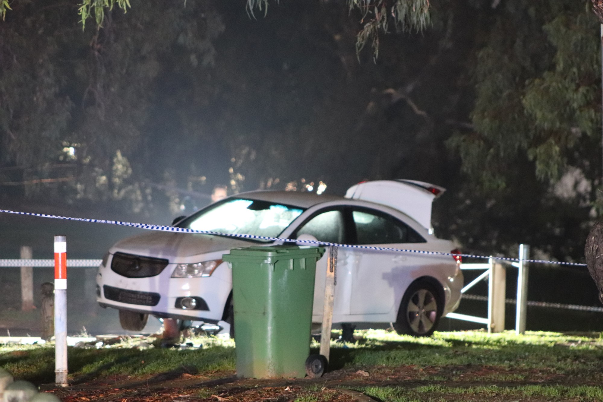 A white car on a grass verge at night with its boot up and a green bin and police tape in the foreground.