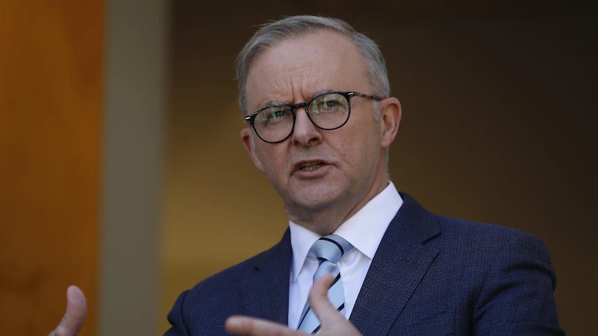 A middle-aged man with short hair and glasses, wears a suit and speaks, gesturing with his hands