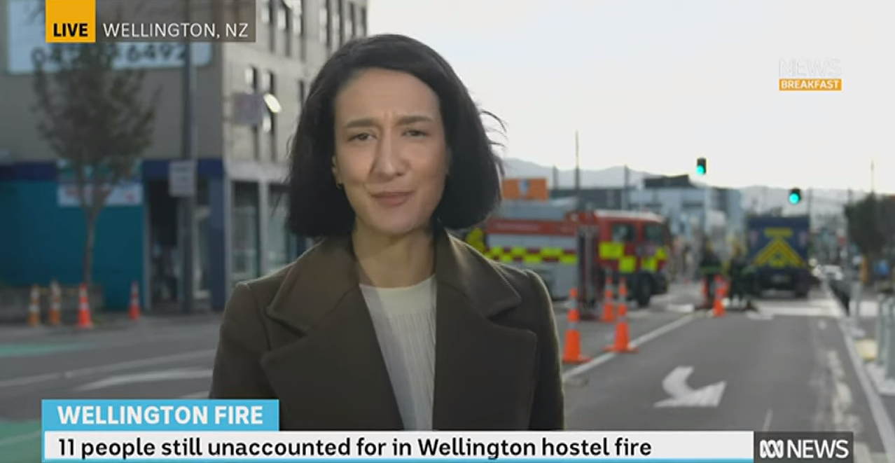 A female reporter with short hair gives an interview from a Wellington street outside a building and near emergency vehicles