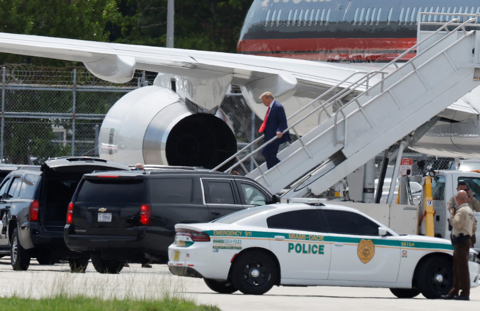 Donald Trump walks down the stairs of an airplane, approaching vehicles