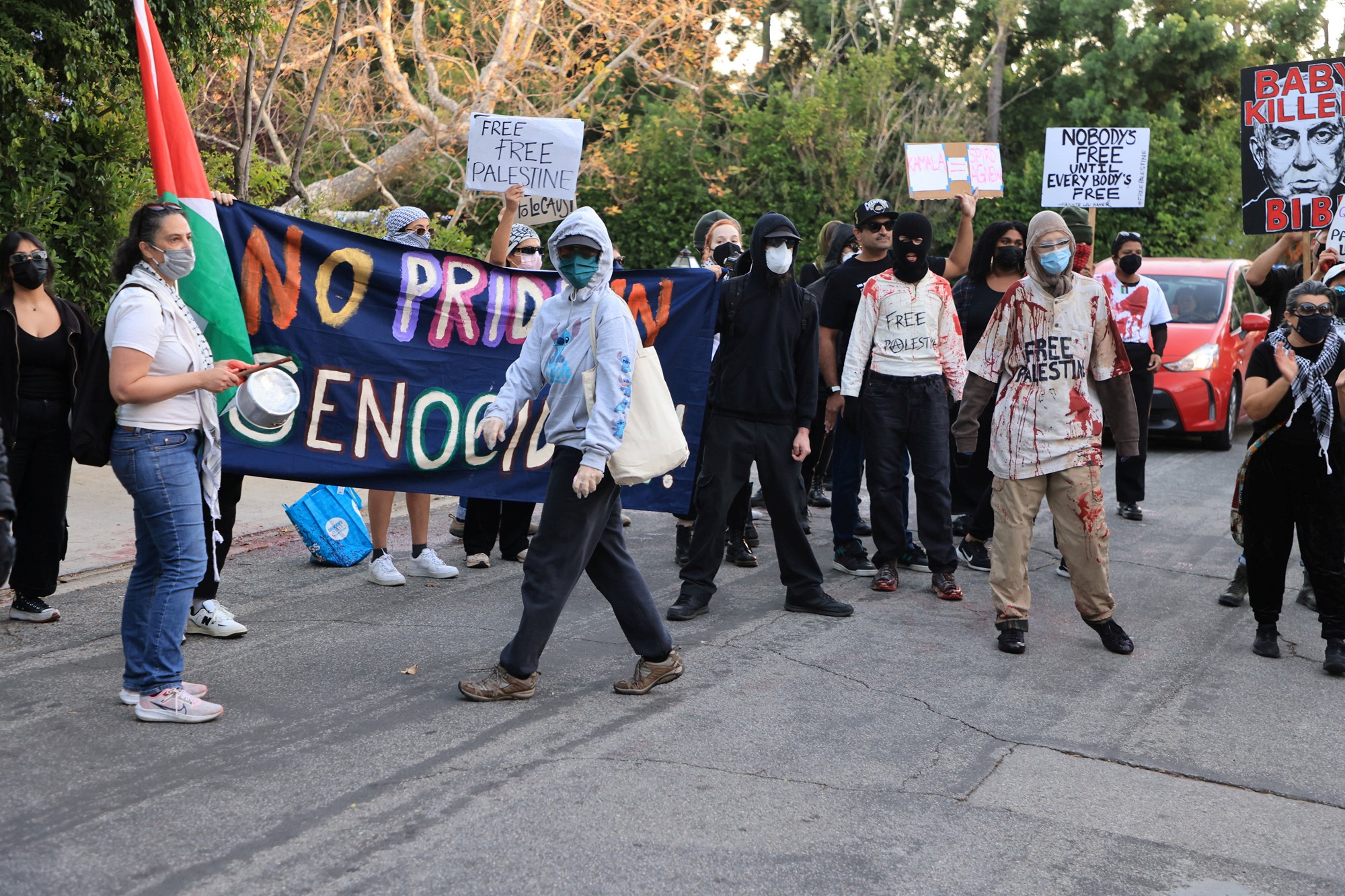 A crowd of protesters with covered faces hold up signs saying 'no pride in genocide'. 