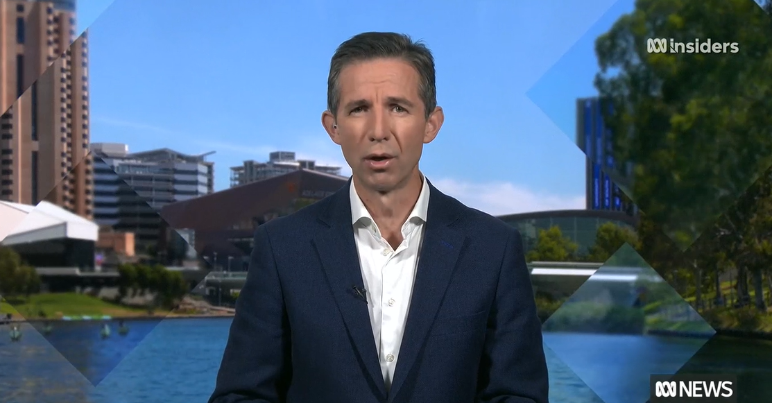 A man in a suit jacket and shirt gives a TV interview in front of a backdrop of Adelaide