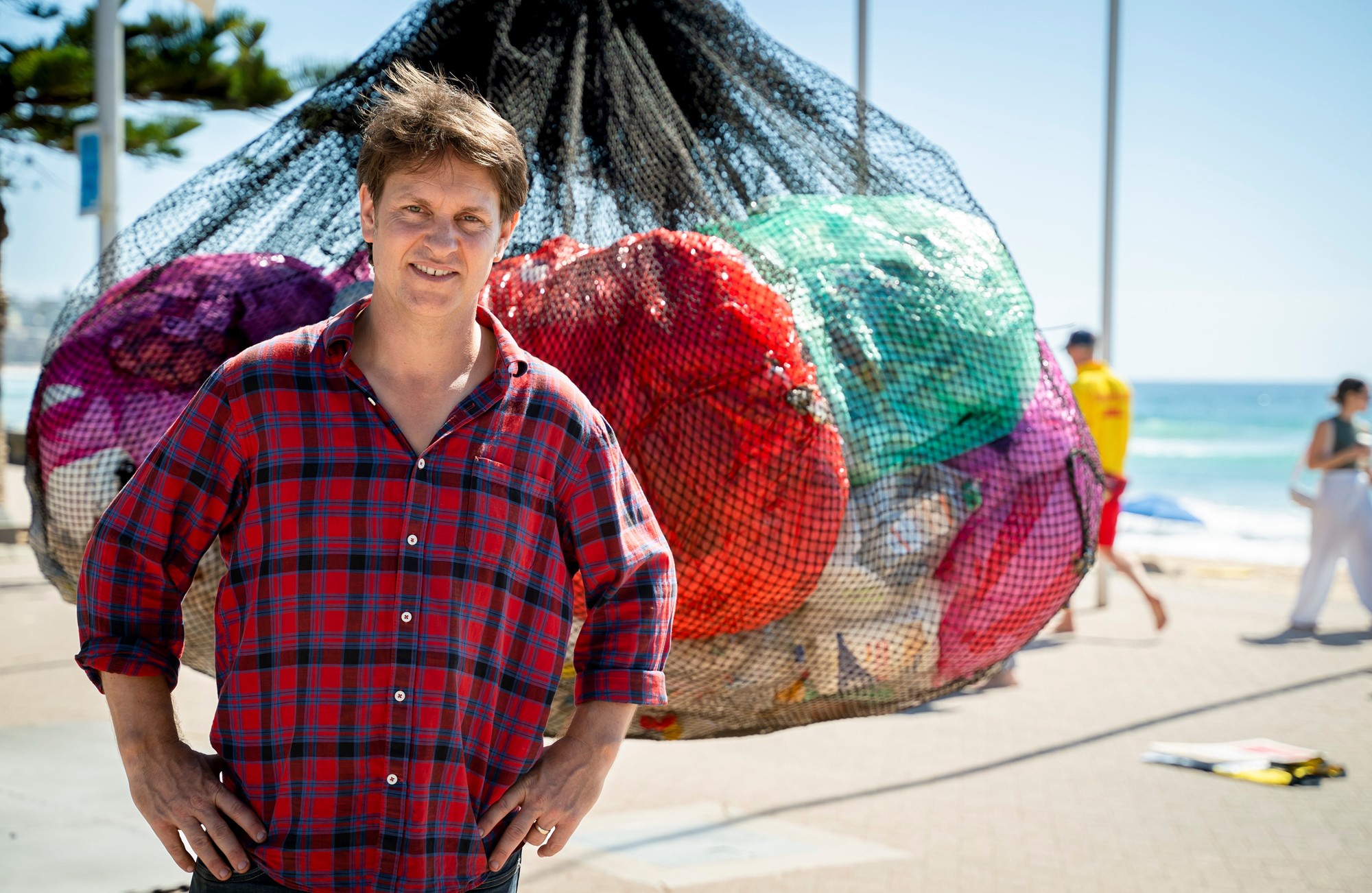 Craig stands with his hands on his hips in front of a black mesh back full of plastic bags that's suspended in the air. The beach is behind him in the background.