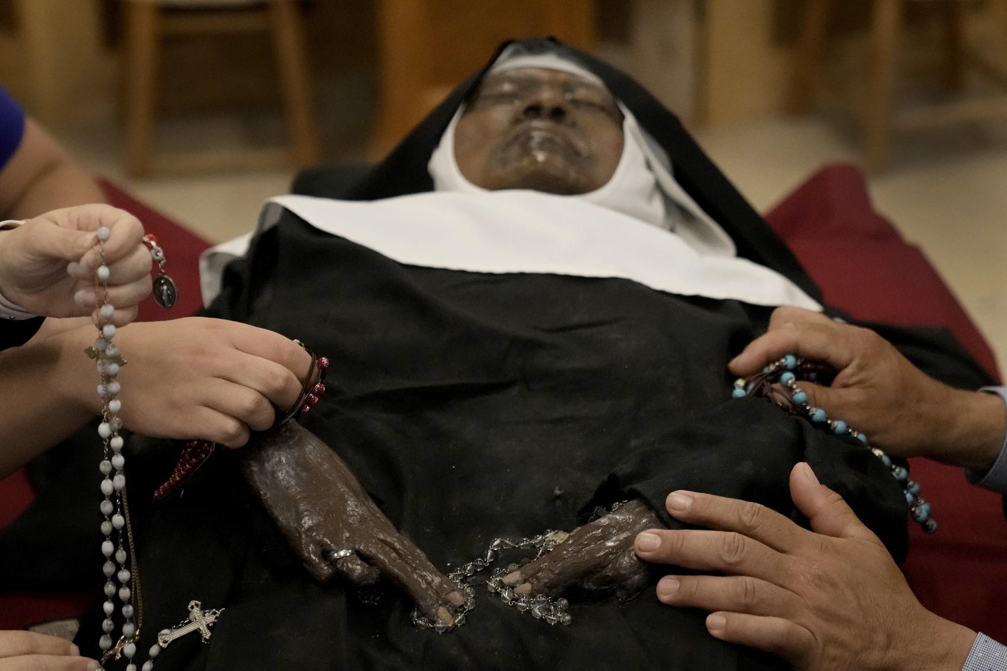 A black nun's body lies in state, with other people's hands touching her and holding rosary beads