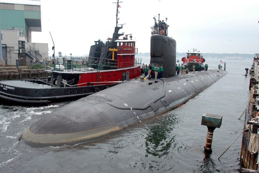 A dark grey submarine  above water, next to a dock.