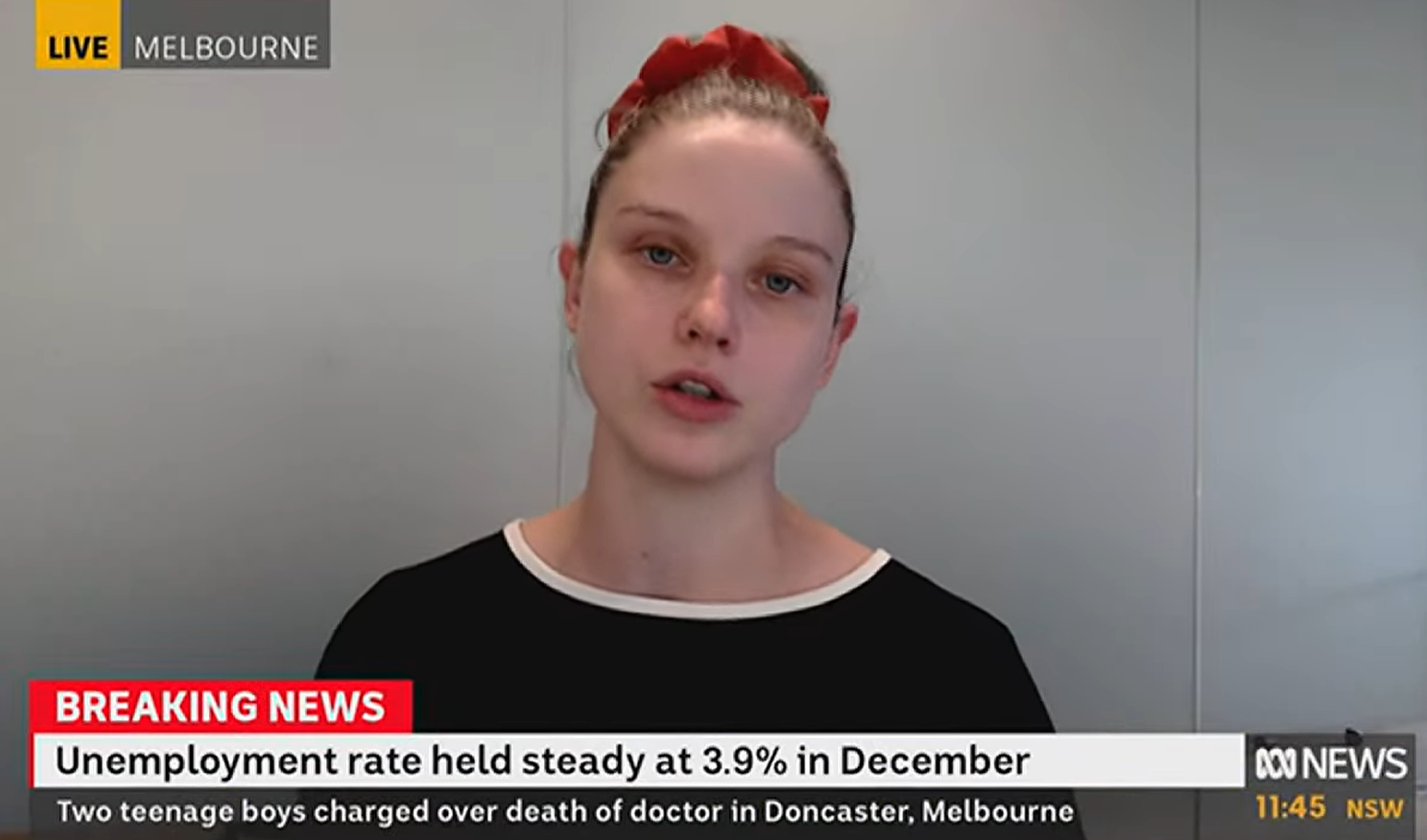 A woman with her hair in a bun secured with a red scrunchie, wearing a black shirt with a thin white collar sits in front of a blank white wall talking. There is a news strap on the bottom of the screen highlighting the unemployment rate is steady at 3.9%, and shows the woman is speaking live in Melbourne.