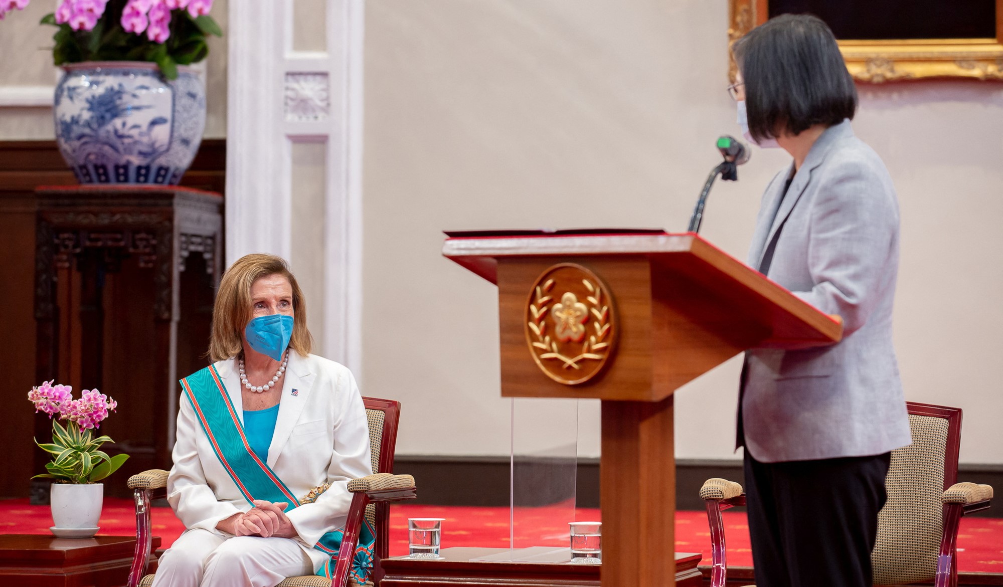 A woman standing behind a lectern looks at another woman who is seated.