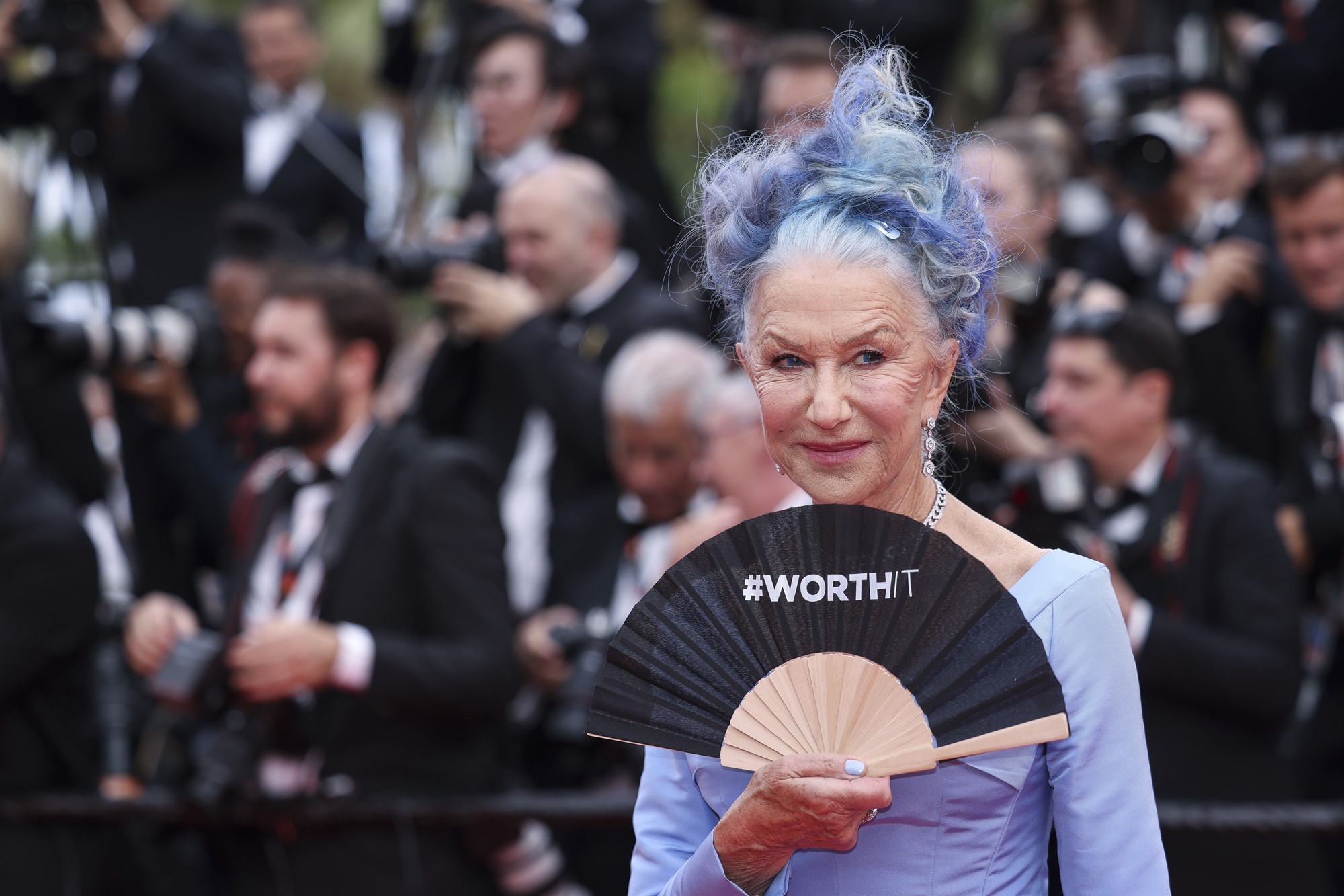 Actor Helen Mirren holding a hand fan with #WorthIt written on it. Photographers are grouped behind her