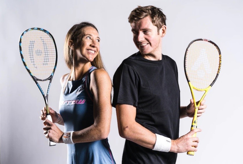 A woman and a man hold racquets back-to-back and look at each other over their shoulders