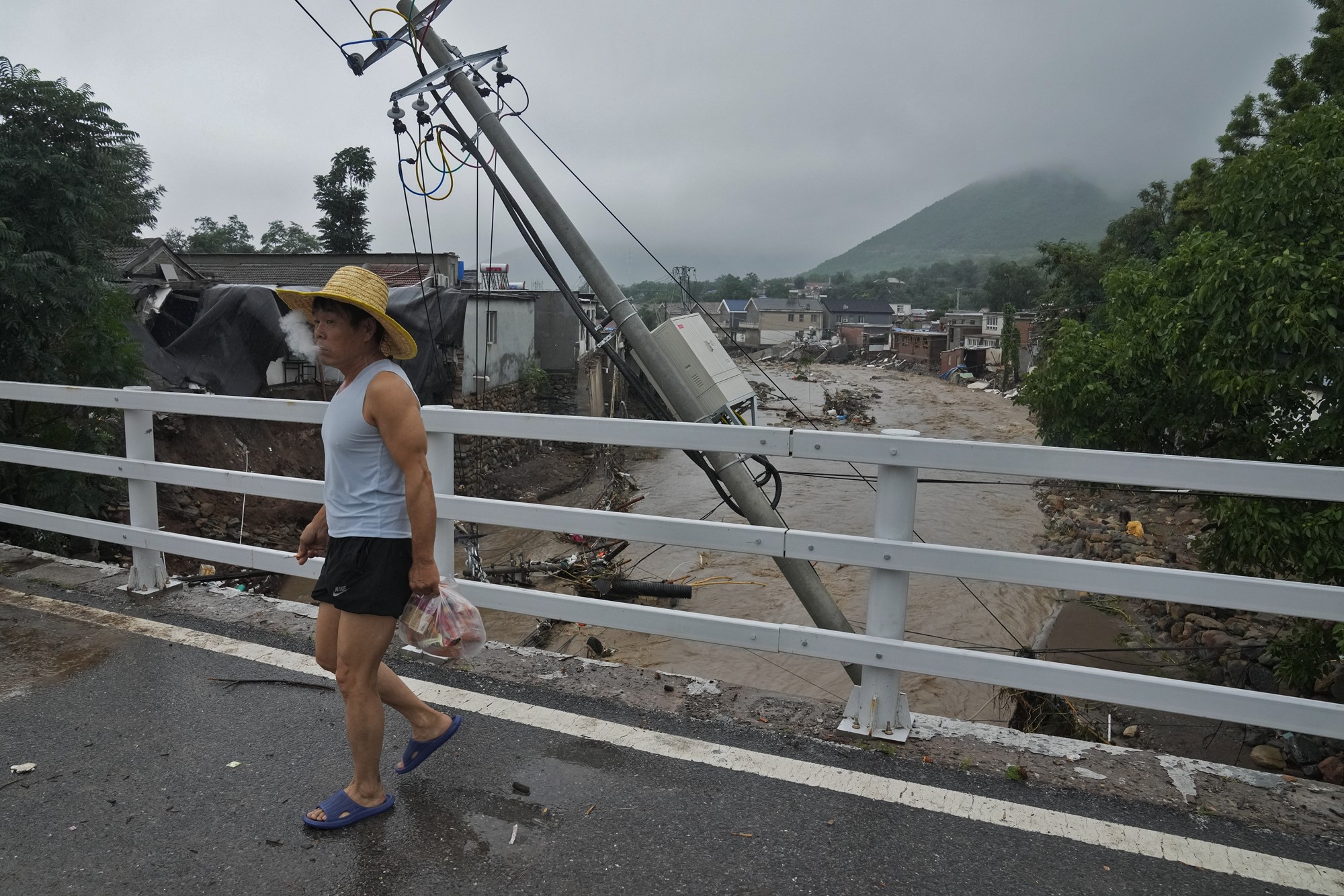 A man in a wide brimmed straw hat and a singlet walks through a village damaged by floodwaters