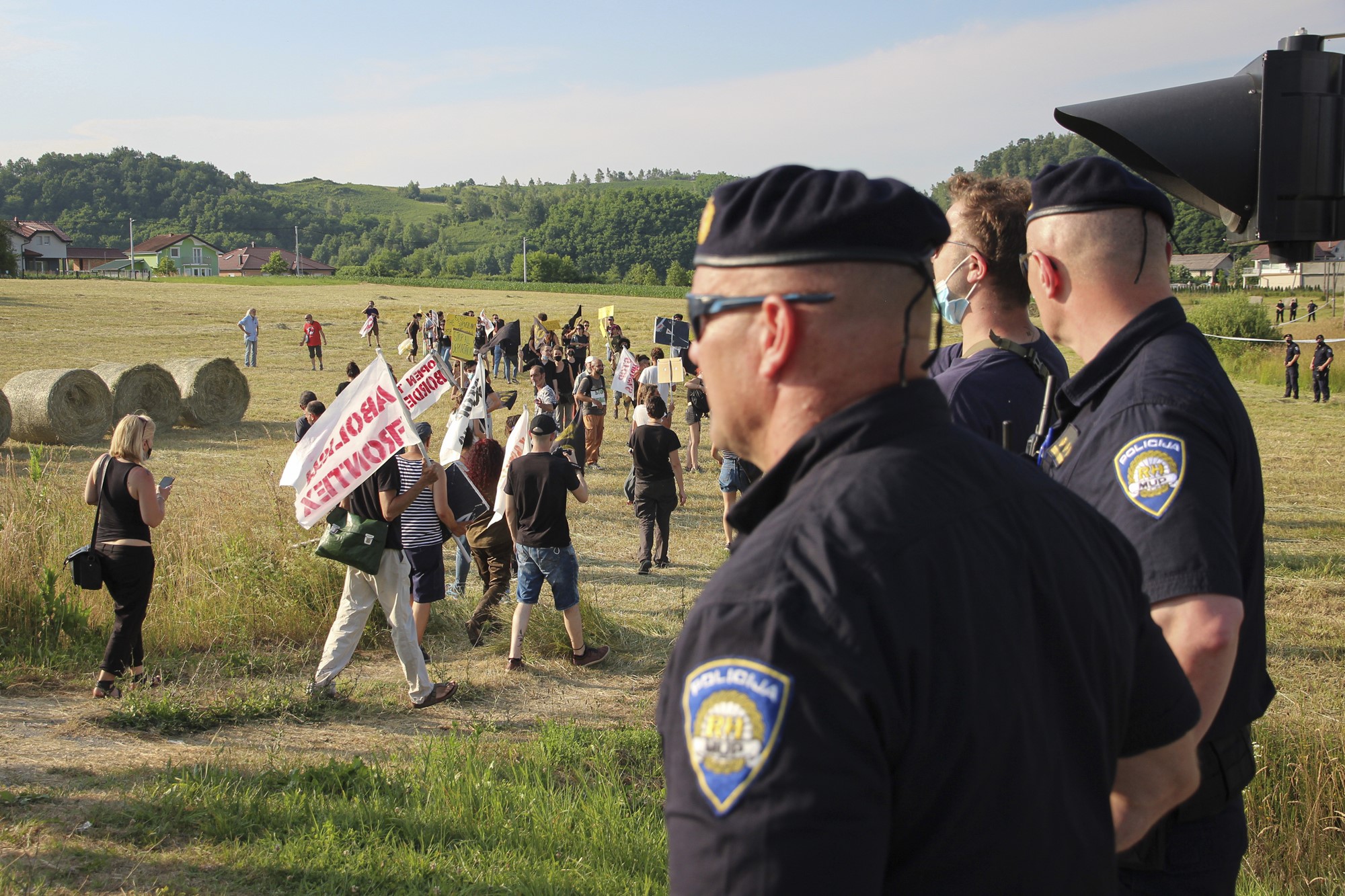 Police stand on a grassy hill watching people bearing flags walk past.