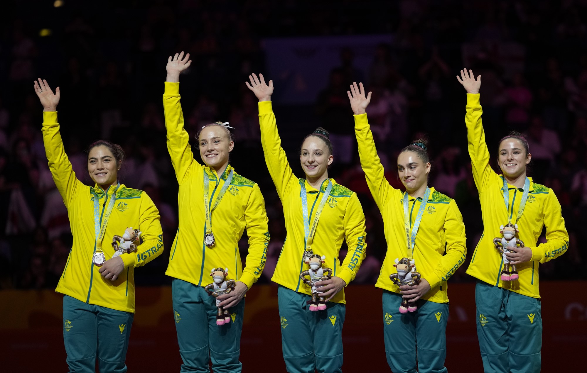 five members of the australian womens gymnastics team pose with medals on a podium smiling and waving
