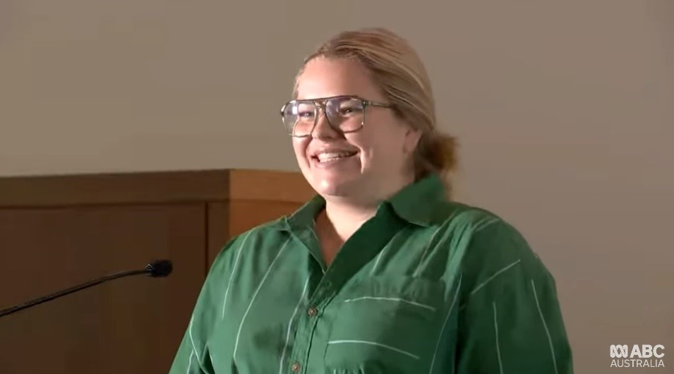 A woman with glasses and a green button-up shirt smiles at a podium.