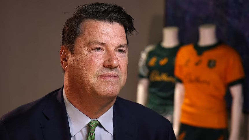 A middle-aged white man in a suit sits in front of two rugby uniforms.