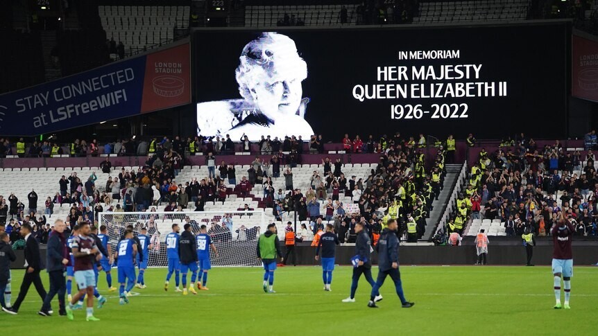 Footballers on a pitch in front of a billboard showing a tribute to Queen Elizabeth II