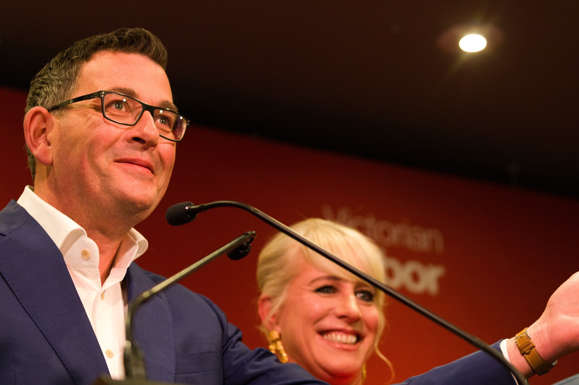 Dan Andrews speaks into a mic with his wife behind him.
