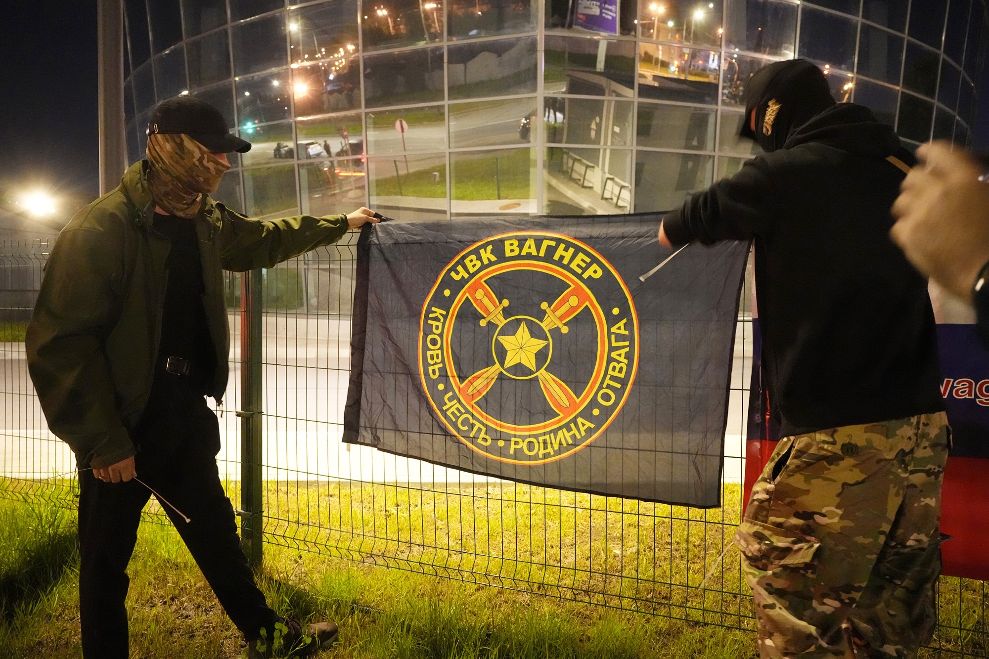 Two men, one wearing camo print pants and the other in black and khaki, both with their faces covered, hang a black flag with Russian writing on it up on a fence