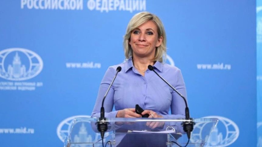 Maria Zakharova stands at a podium in front of a blue backdrop. 