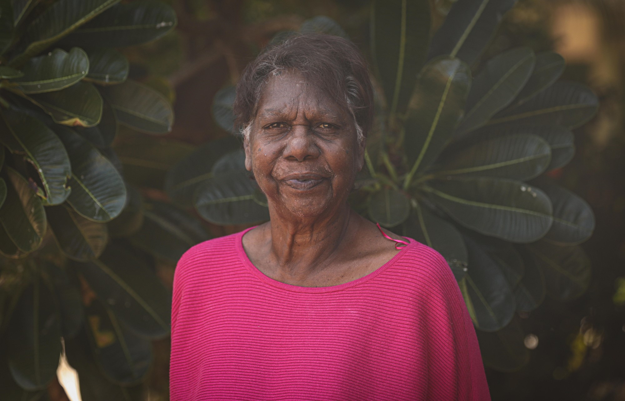 An Aboriginal woman stands in front of a deep green tree.
