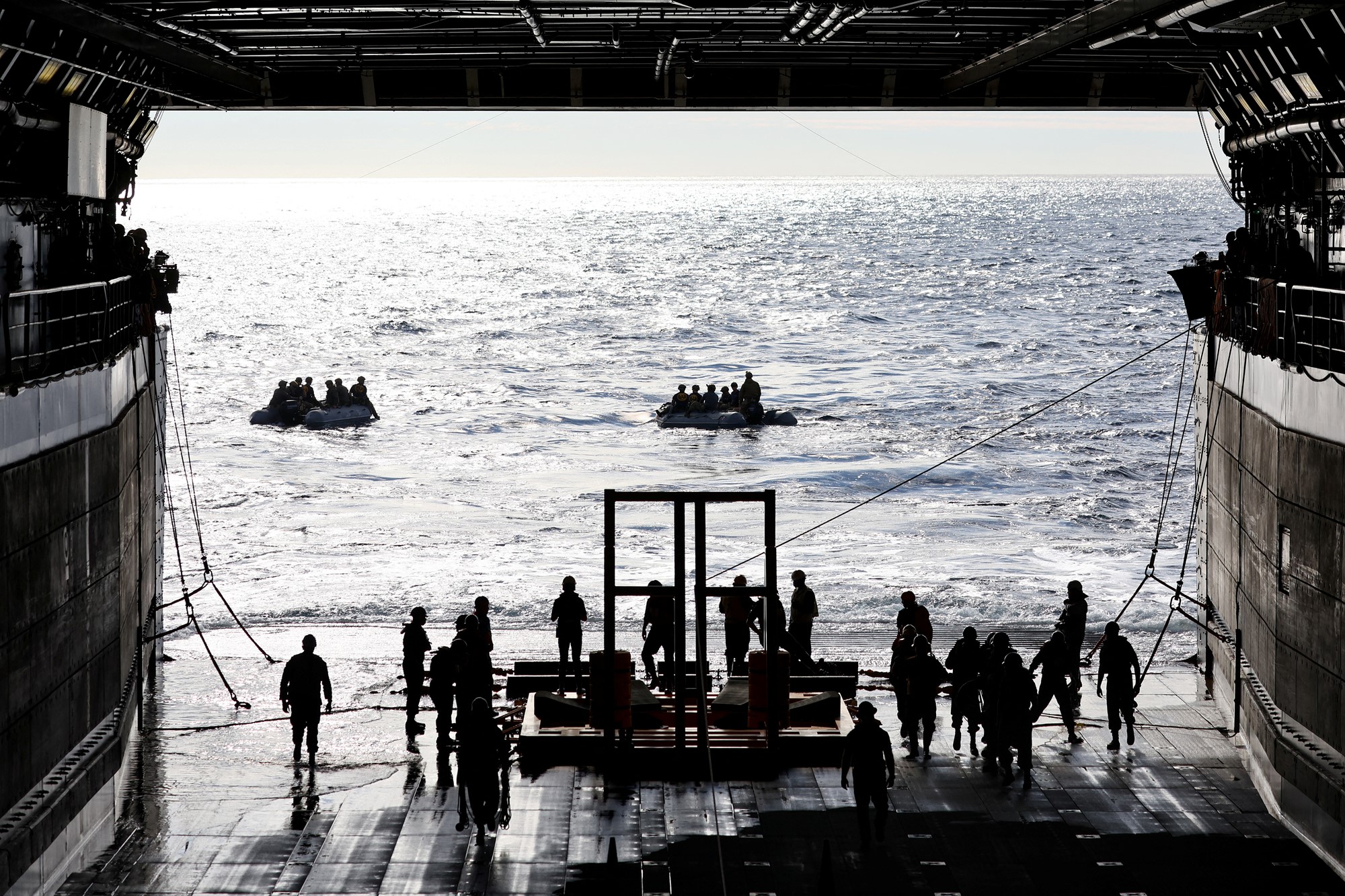 US Navy men lead a boat into th ocean out of a ship hangar. 