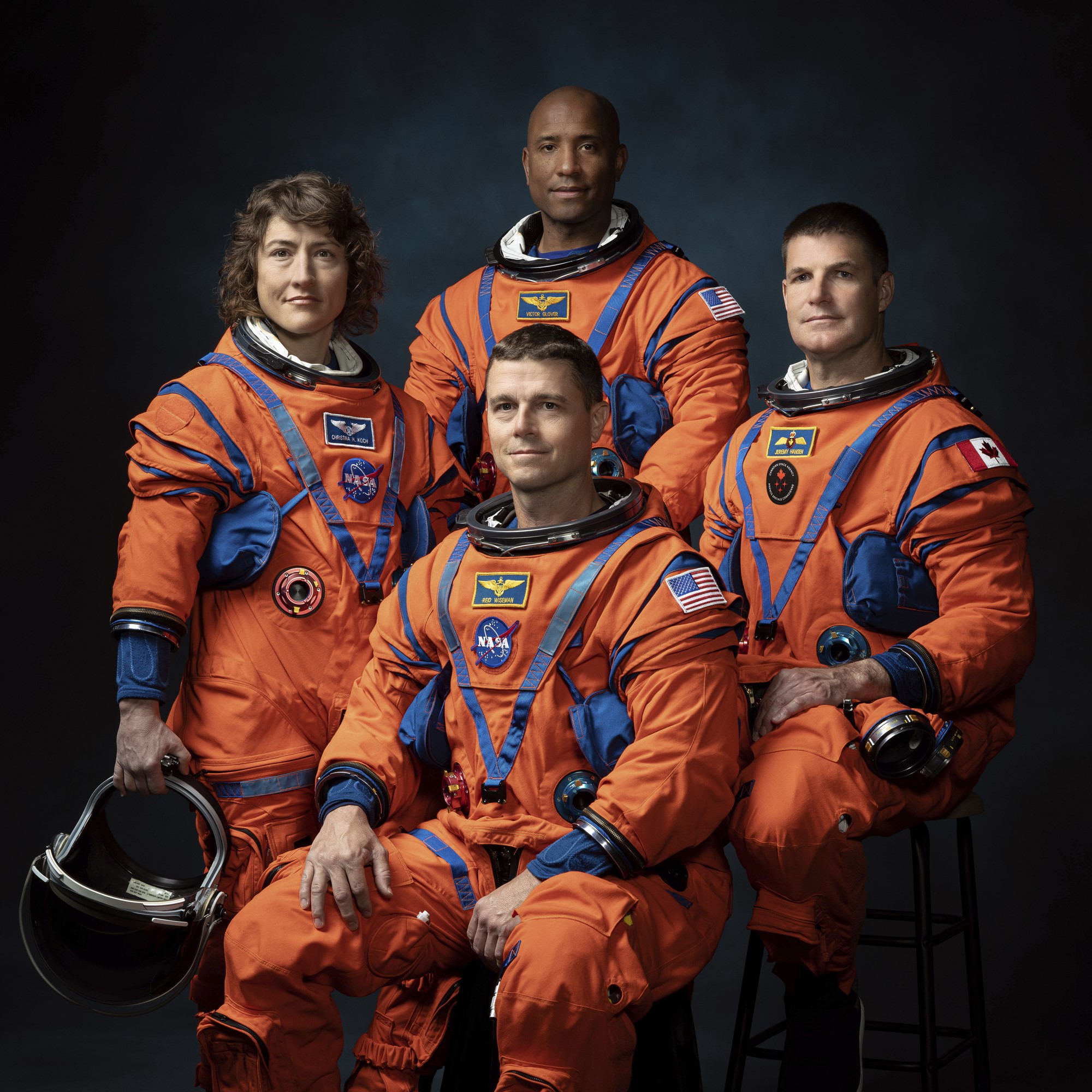 A photo of four astronauts, three standing and one sitting, all in their NASA spacesuits and smiling. One holds a helmet.