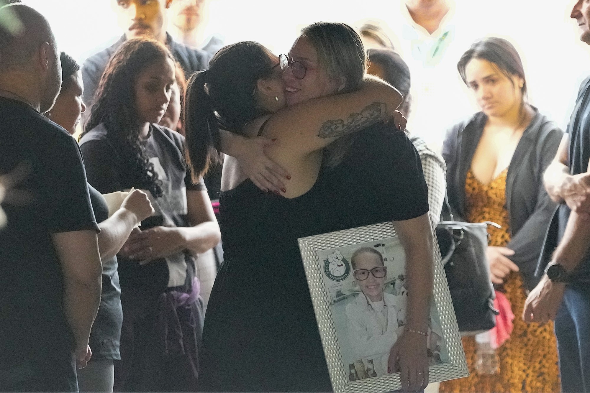 Two women hug amid a crowd, one holding a framed portrait of a little girl.