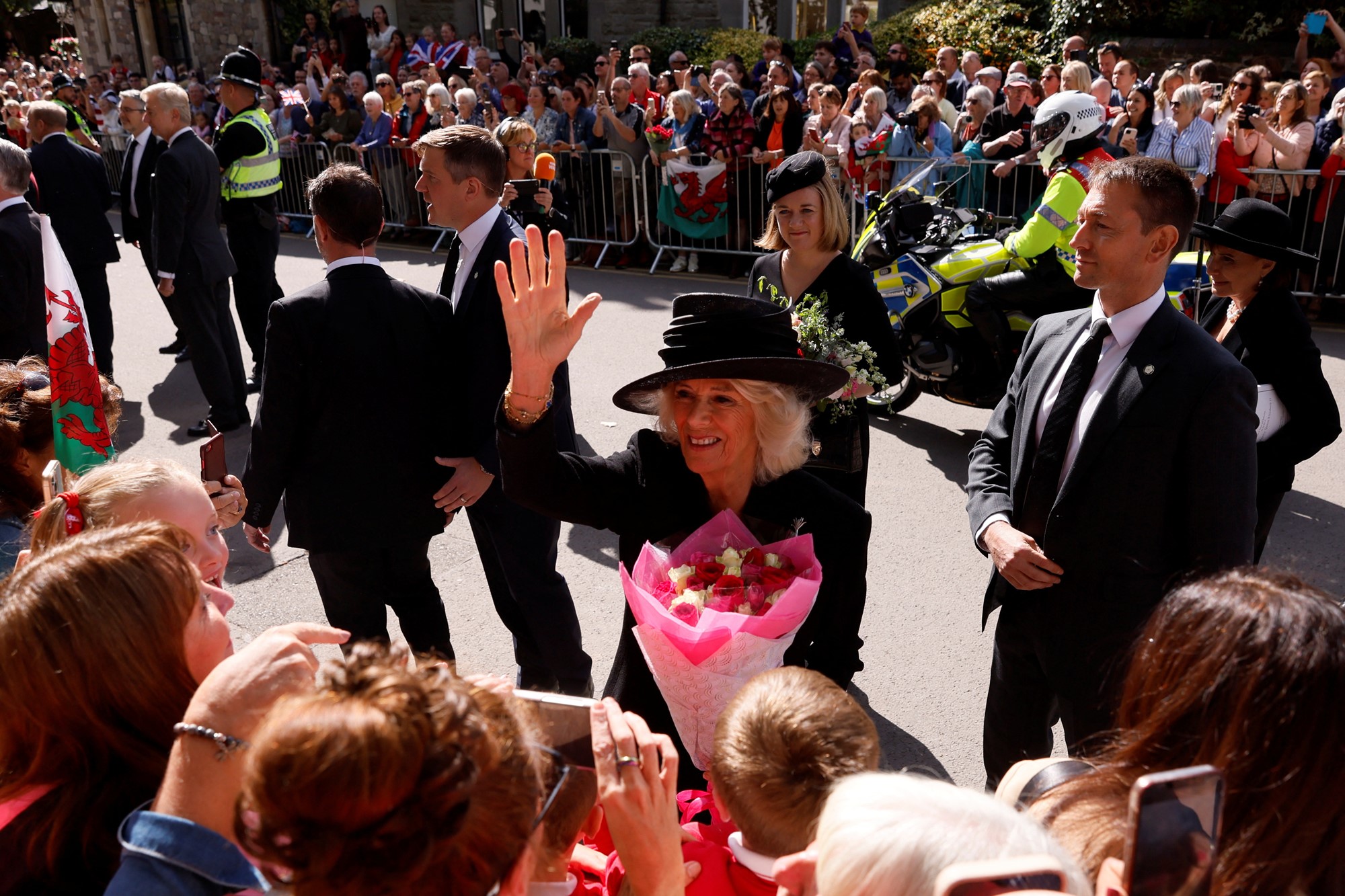 Camilla waves at crowd as a holds pink flowers.