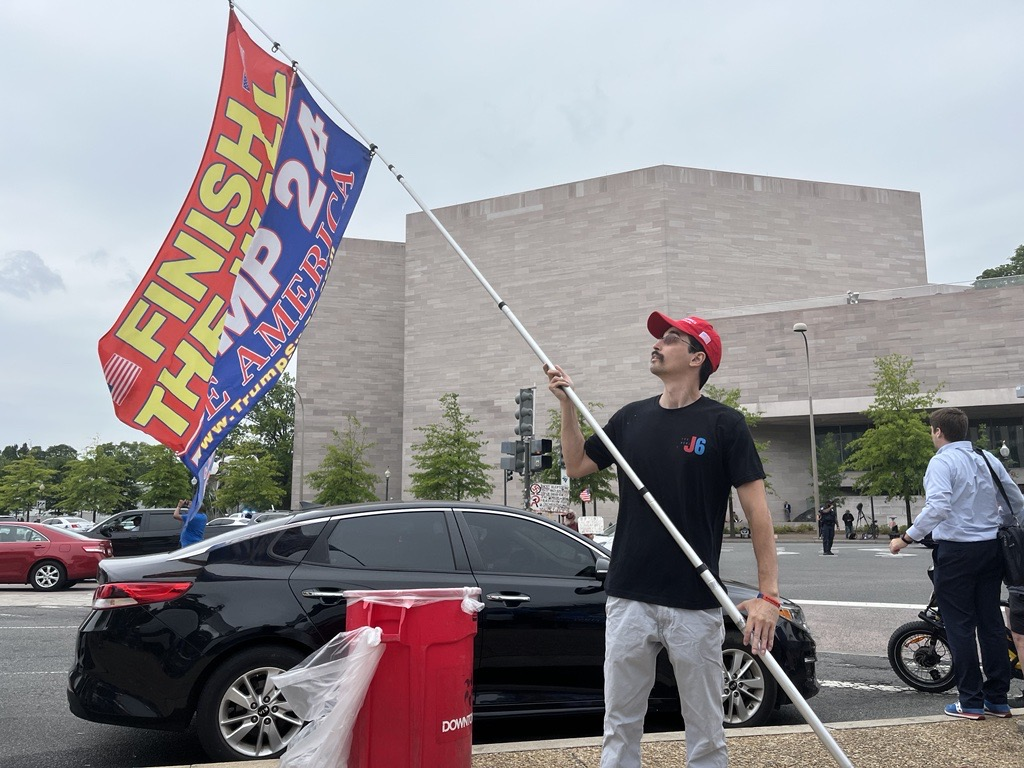 A man wearing a black T-shirt and red cap waves a MAGA flag