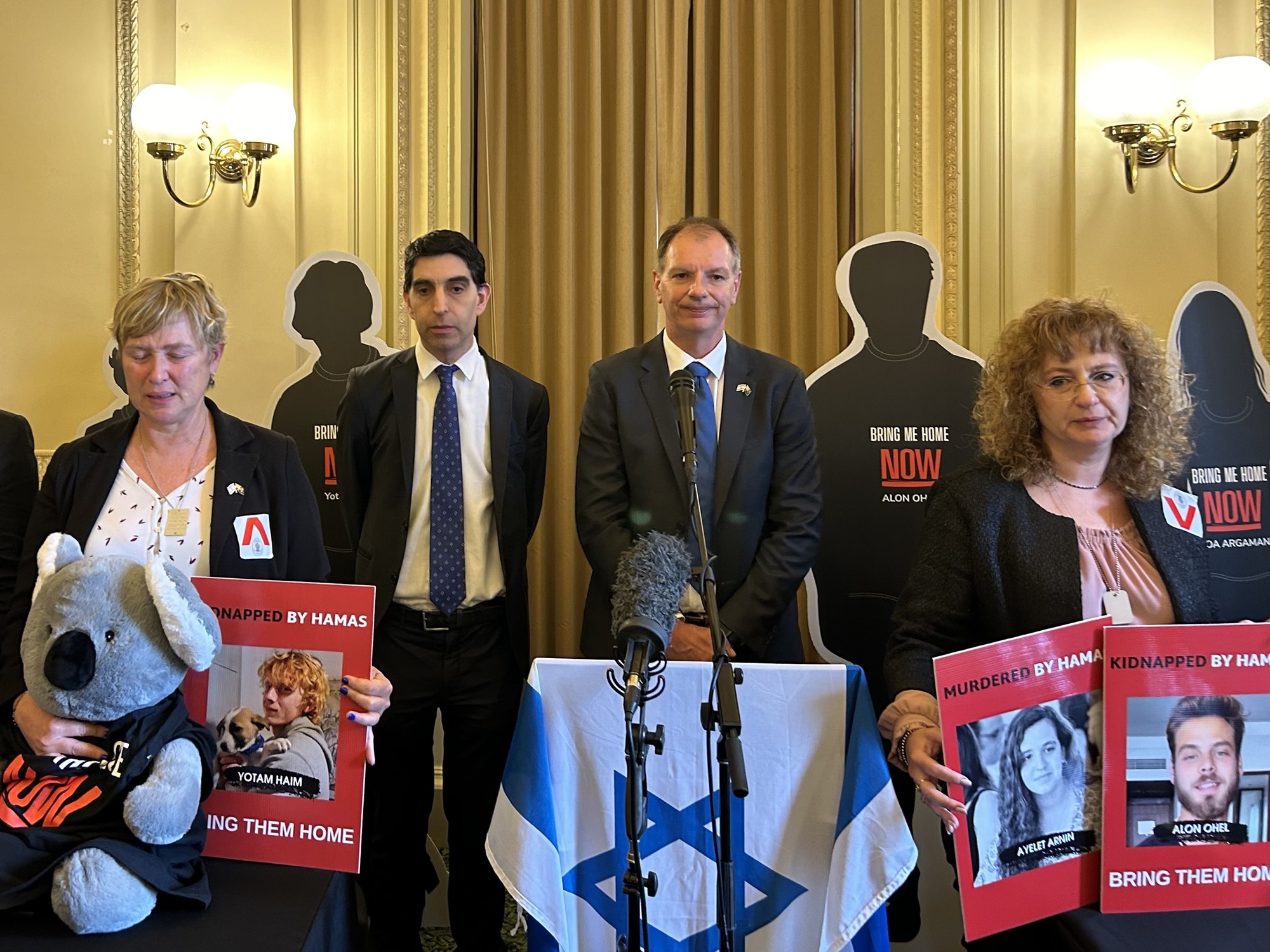 Four people wearing black and holding signs standing near a podium with israeli flags and carboard cutout silhouettes