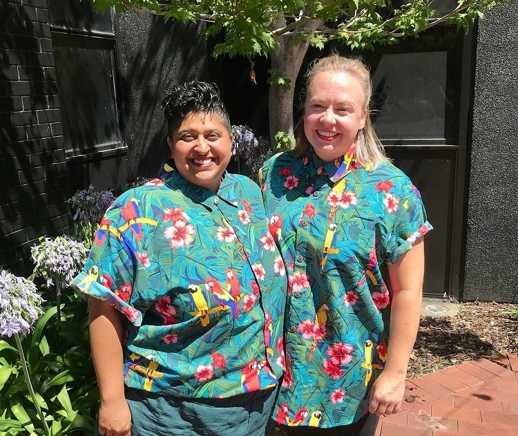Two women in matching parrot t-shirts stand together smiling. One is shorted with brown hair and one is taller with blonde hair.
