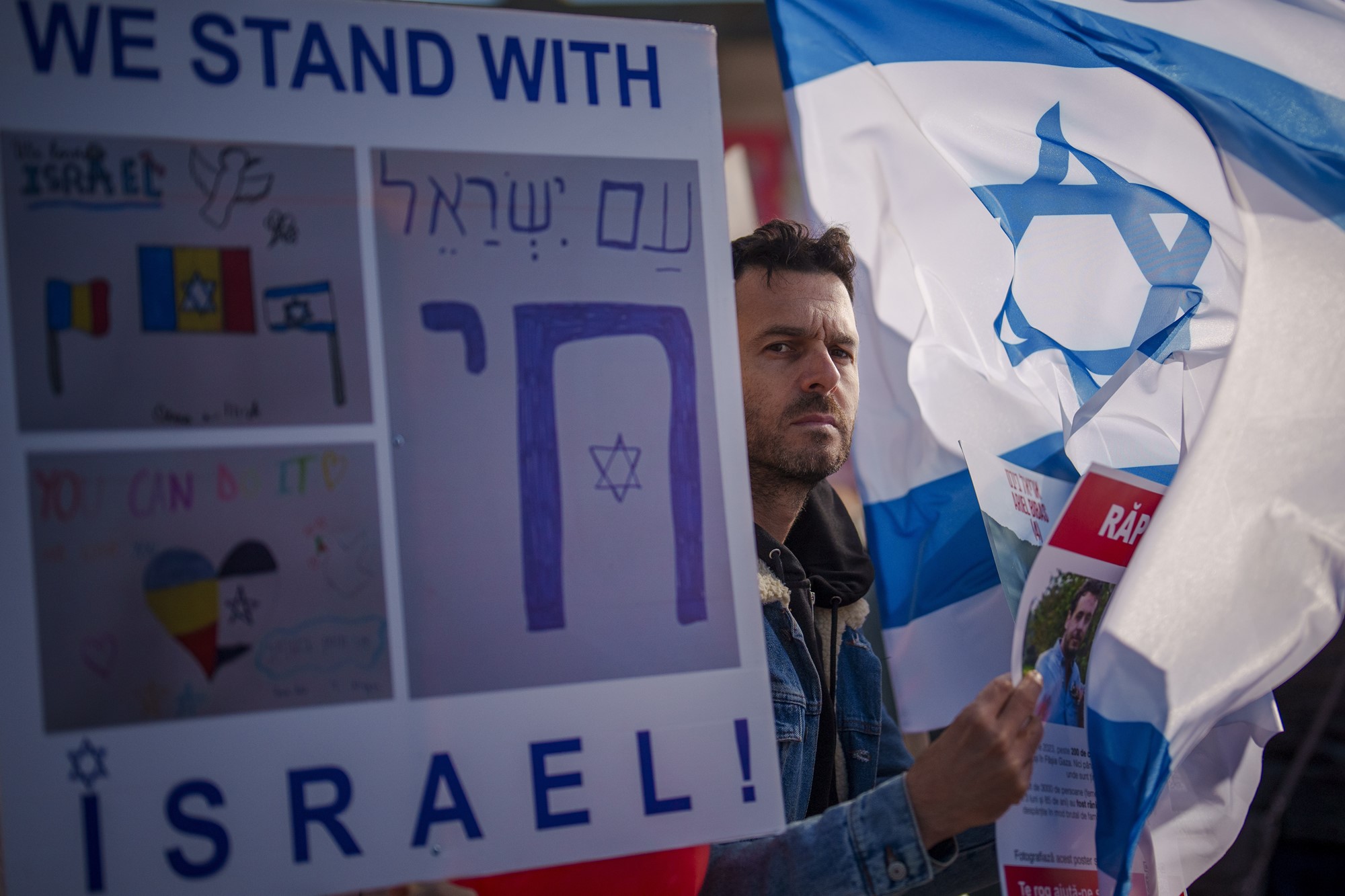 Man stands with missing poster and Israel flag in hand next to sign saying we stand with israel. 