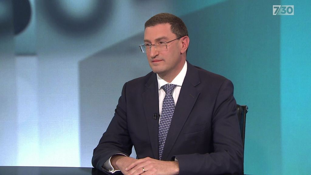 A man sits at a desk wearing a suit and glasses.