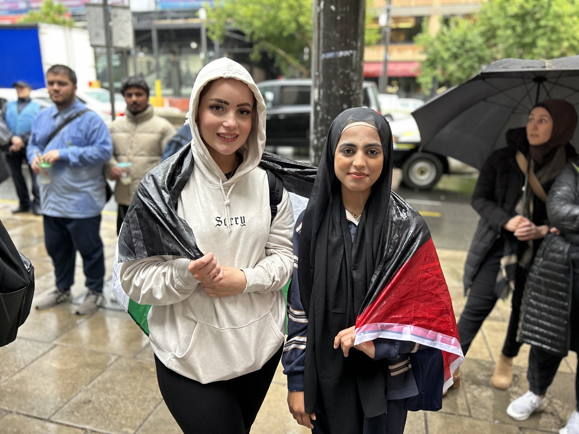 Two girls are pictured, they look rained on and are wearing a Palestinian flag over their shoulders.