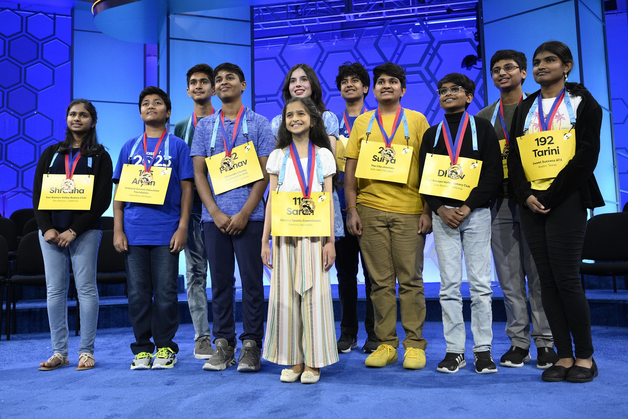 A group of 11 children standing together on a stage, with large nametags and medals around their necks