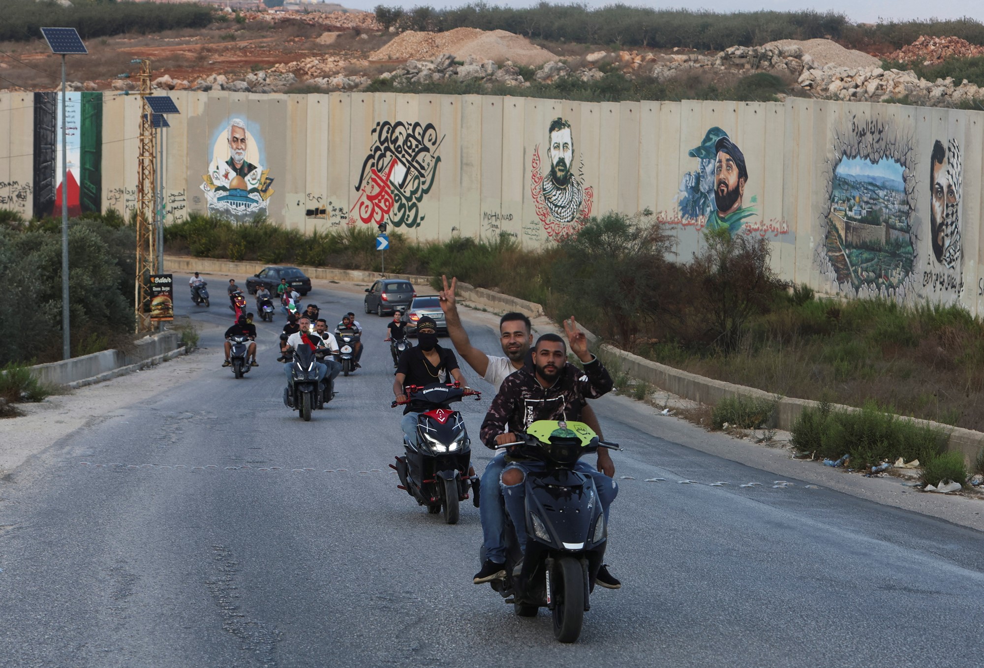 Several men on motorbikes driving on a road, making the peace symbol.