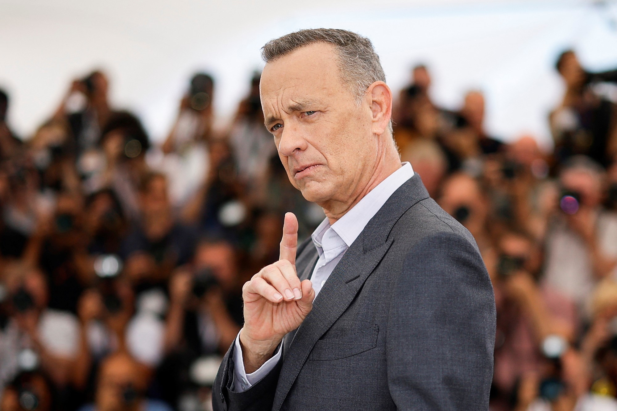 Tom Hanks raises a finger while on a red carpet in front of photographers.